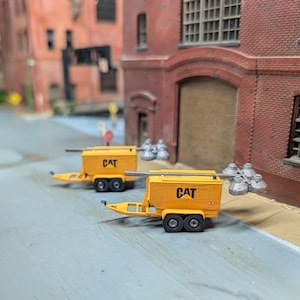 May include: Two yellow CAT construction trailers with black wheels are parked on a gray street. The trailers have a rectangular shape and are attached to a yellow hitch. The background features brick buildings and a street scene.