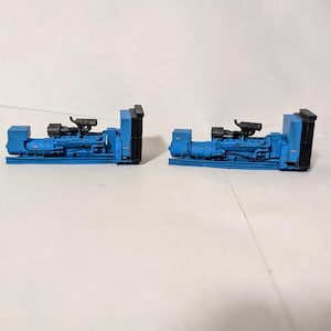 May include: Two blue industrial generators with black components are displayed on a white surface. The generators have detailed piping and exhaust systems. The overall design suggests a model or miniature representation of heavy machinery.
