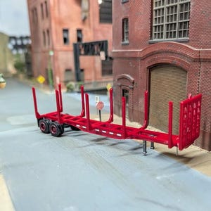 May include: A red model logging trailer with vertical supports and a rear gate. The trailer has three sets of black wheels. It is parked on a grey surface. The background features brick buildings and a street.