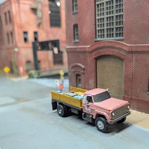May include: A miniature pink flatbed truck with a yellow bed, parked on a gray street. The truck is in front of a brick building. The truck bed contains small, rectangular objects.