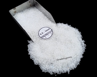 May include: A close-up shot of a pile of small, clear, granular material, possibly gemstones or decorative elements, spilling from a metal scoop. A circular scale indicates a size range of 0.5-1mm. The background is black.