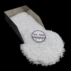 May include: A close-up shot of a pile of small, clear, crystalline particles, possibly a mineral or chemical compound, spilling from a metal scoop. A circular label indicates a size range of 0.5-1mm.