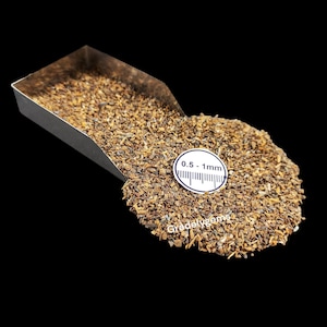 Puede incluir: Primer plano de un montón de material granular pequeño y marrón, posiblemente piedras preciosas o minerales, que se derrama de una cuchara de metal. Una etiqueta circular blanca indica un tamaño de 0,5 - 1 mm. El fondo es negro.