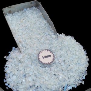 May include: A pile of light blue, crushed glass pieces, with a metal scoop pouring them out. A small, circular label reads "1-3mm". The glass pieces are small and irregular in shape, with a slightly translucent appearance. The background is black.