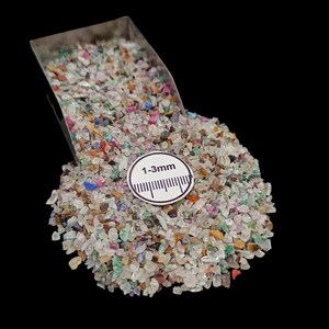 Pode incluir: Uma pilha de pequenas lascas de pedras preciosas coloridas com uma concha de metal. As lascas são uma mistura de transparente, rosa, azul, verde e laranja. Um rótulo circular branco indica um tamanho de 1-3 mm.