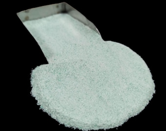 May include: A close-up shot of a small, stainless steel scoop filled with a pile of light green, granular material. The material has a slightly textured appearance and is set against a solid black background.