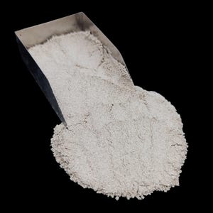 Pode incluir: Uma pá de metal despejando um monte de pó fino, cinza claro, contra um fundo preto. O pó parece granular e tem uma superfície ligeiramente texturizada. A pá é feita de metal brilhante de cor prateada.