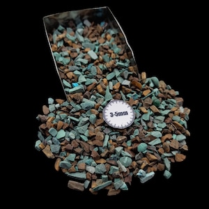 May include: A pile of small, irregularly shaped stones in shades of teal and brown, with a metal scoop partially visible at the top. A white circular tag with the text "3-5mm" rests on the stones. The stones are scattered on a black background.