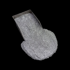 Puede incluir: Primer plano de una pequeña pala metálica rectangular llena de finos gránulos plateados. Los gránulos se derraman sobre un fondo negro, creando un efecto texturizado y brillante. La imagen está bien iluminada, resaltando el brillo metálico.