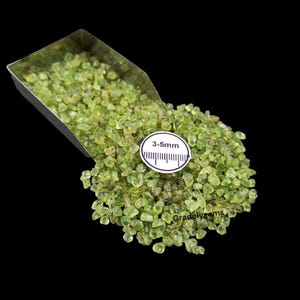 May include: A pile of small, light green peridot gemstones, with a size range of 3-5mm, spilling from a metal scoop. A white circular tag with the size measurement is visible. The background is black.