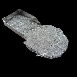 May include: A close-up shot of a pile of dark gray, granular material, possibly a mineral or gemstone, spilling from a small, silver metal tray. The material has a textured surface with small, reflective particles. The background is black.