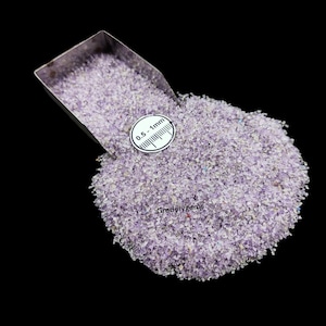 Puede incluir: Un montón de pequeños fragmentos de piedras preciosas de color púrpura claro con una cuchara de metal. Un indicador circular blanco indica un tamaño de 0,5 a 1 mm. El texto "Gradelygems" es visible en la parte inferior.