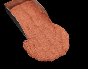 May include: A metal scoop holding a pile of fine, reddish-brown powder. The powder spills out onto a black surface. The scoop is angled, showing the texture of the powder.