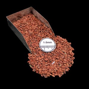 Op de afbeelding: Een metalen schep gevuld met kleine, roodbruine grind. Een wit rond label met de tekst "1-3mm" en een millimeterliniaal is op de grind geplaatst. Het grind is verspreid op een zwarte achtergrond.
