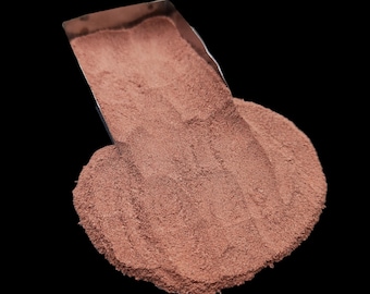 May include: A pile of fine, reddish-brown powder is shown against a black background. A stainless steel scoop is partially visible, holding some of the powder. The powder is spilling out of the scoop, creating a small mound.