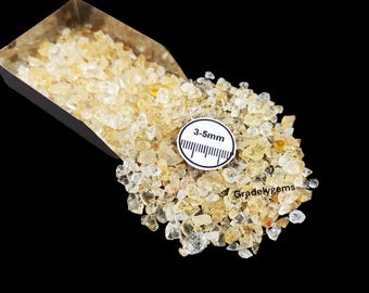 May include: A pile of small, light yellow-brown gemstones, with a scoop partially visible at the top left. A white circular tag indicates the stones are 3-5mm in size. The background is black, highlighting the gems.