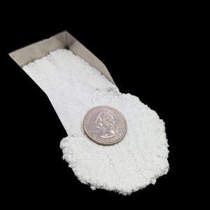 May include: A close-up shot shows a pile of white, crystalline material next to a U.S. quarter coin. The material is in a small, silver metal scoop. The background is solid black.