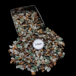 Puede incluir: Un montón de pequeñas piedras de colores en tonos verdes, marrones y blancos. Una cuchara de metal es parcialmente visible, vertiendo las piedras. Una pequeña etiqueta circular indica un tamaño de 3-5 mm.