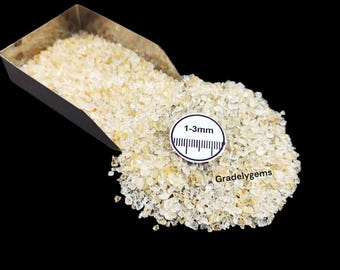 May include: A pile of small, light yellow gemstones, possibly citrine, spilling from a metal scoop. A white circular gauge with the text "1-3mm" and a ruler is placed on the gemstones. The background is black.