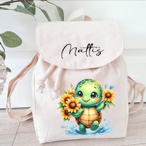 Könnte beinhalten: Ein beigefarbener Canvas-Rucksack mit Kordelzug. Der Rucksack hat eine niedliche Comic-Schildkröte mit Sonnenblumen und den Namen "Mattis" auf der Vorderseite.