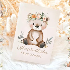 Könnte beinhalten: Ein cremefarbenes Babybuch mit einer Aquarellillustration eines Teddybären mit Blumenkranz. Das Buch ist von anderen Tierillustrationen und dem Text "Ultraschallbilder Marie Sommer" umgeben.