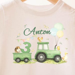 Könnte beinhalten: Weißes T-Shirt mit einem grünen Traktor, der einen Wagen mit der Zahl 3 und einem Hund zieht. Das Shirt hat den Namen "Anton", Ballons und Illustrationen von Hoftieren.