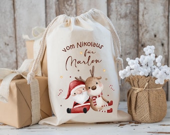 May include: A cream-colored drawstring gift bag with a Santa Claus and reindeer illustration. The bag has the text "vom Nikolaus für Marlon" in brown script. Presents wrapped in brown paper and a burlap vase with white flowers are in the background.