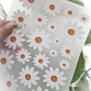 Könnte beinhalten: Ein Bogen mit weißen Gänseblümchen-Aufklebern mit orange-braunen Zentren. Die Aufkleber sind in verschiedenen Größen auf einem durchscheinenden Hintergrund angeordnet. Das Bild ist aus der Nähe aufgenommen und hebt das florale Design hervor.