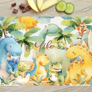 Könnte beinhalten: Ein farbenfroher Platzdeckchen mit einem skurrilen Dinosaurier-Motiv. Das Platzdeckchen hat einen weißen Hintergrund mit grünem Laub und verschiedenen Dinosauriern in Blau, Gelb und Grün. Der Text "Milo" ist in einer verspielten Schriftart auf dem Platzdeckchen geschrieben.