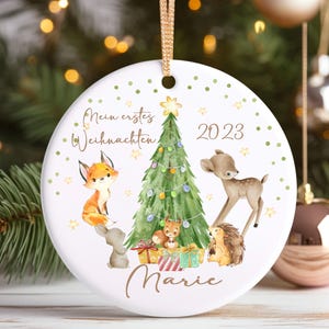 Könnte beinhalten: Ein weißer Weihnachtsbaumschmuck aus Keramik mit goldenem Band. Der Schmuck zeigt einen Weihnachtsbaum, Waldtiere und den Text "Mein erstes Weihnachten 2023 Marie". Das Design umfasst einen Fuchs, ein Reh, ein Eichhörnchen, einen Igel und Geschenke.