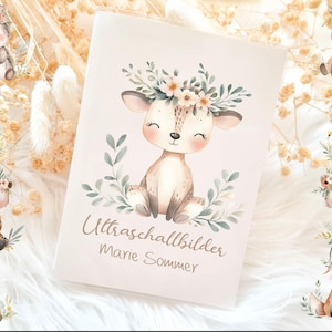 Könnte beinhalten: Ein cremefarbenes Babybuch mit einer Aquarellillustration eines Rehs mit Blumenkranz. Der Text "Ultraschallbilder Marie Sommer" ist auf dem Einband aufgedruckt. Um das Buch herum befinden sich Illustrationen von Tieren wie Igel und Pinguin.