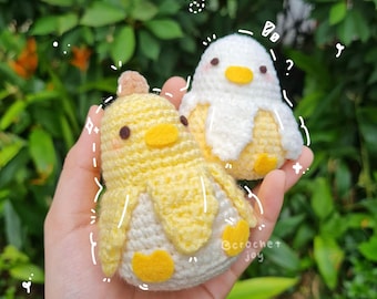 May include: Two crocheted chick plushies, one yellow and one white, with black eyes and yellow beaks. The plushies are held in a hand against a green background.