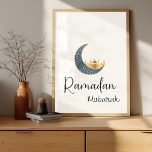 Puede incluir: Un cuadro enmarcado con las palabras "Ramadan Mubarak". La obra presenta una luna creciente con un patrón azul y dorado, y una silueta dorada de mezquita. El marco es de madera clara.