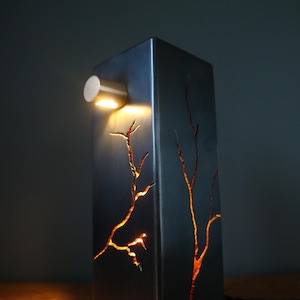 May include: A modern, rectangular table lamp with a dark gray metal finish. The lamp features a small, round light fixture and intricate, tree-like cutouts that glow with warm light. The lamp is on a wooden surface.