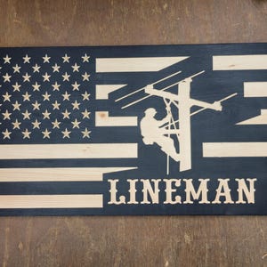 May include: A wooden sign with an American flag design and a silhouette of a lineman working on a power pole. The text "LINEMAN" is below the image.