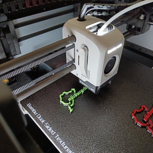 Könnte beinhalten: Ein weißer 3D-Drucker mit einem schwarzen und grauen, strukturierten Druckbett. Der Drucker druckt ein grünes Plastikmodell der Nürburgring-Rennstrecke. Das Druckbett ist mit "Bambu Lab Dual Sided Textured" beschriftet.