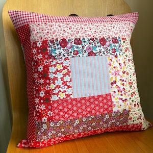 May include: A square patchwork cushion featuring floral and geometric patterns in red, white, and blue. The cushion has a gingham top and a red polka dot border. The cushion is approximately 40 cm square.