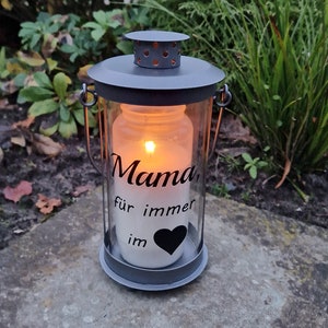 Könnte beinhalten: Eine graue Metall-Laterne mit einem Glasgefäß im Inneren. Das Gefäß enthält eine weiße Kerze mit einem schwarzen Herzen und dem Text "Mama für immer im" darauf.