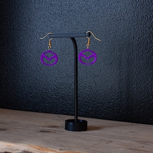 May include: A pair of purple earrings with a geometric design, hanging from a black stand on a wooden surface.