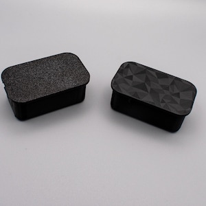 May include: Two black rectangular boxes with different surface textures. One box has a smooth, textured surface, while the other has a geometric pattern.