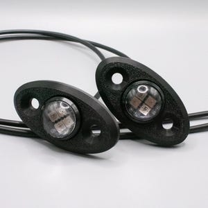 May include: Two black LED lights with oval housings and two mounting holes each. The lights are connected by black wires. The lights have a clear lens and are designed for automotive use.