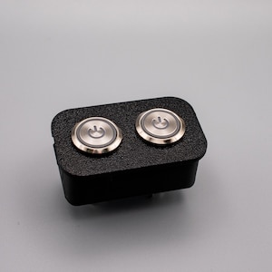 Könnte beinhalten: Eine schwarze rechteckige Box mit zwei runden, silberfarbenen Knöpfen. Jeder Knopf hat ein erhabenes Stromsymbol.