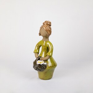 May include: A ceramic figurine of a woman in a green dress, holding a basket of white daisies. The figure has brown hair and a neutral skin tone. The basket is dark brown with a diamond pattern.