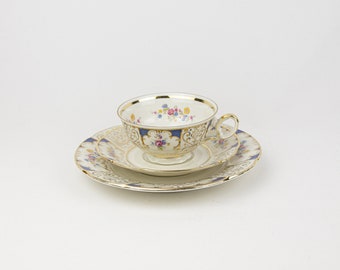 May include: A white teacup and saucer set with gold trim and floral designs. The cup has a blue band with gold accents and the saucer has a gold rim.