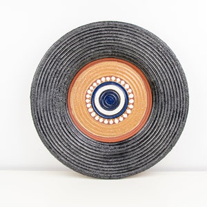Könnte beinhalten: Ein schwarzer und brauner Keramik-Wandteller mit konzentrischem Kreisdesign. Der mittlere Kreis ist blau und weiß mit einem kleineren blauen Kreis in der Mitte. Der äußere Kreis ist schwarz mit einer strukturierten Oberfläche.