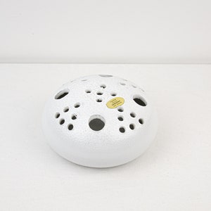 May include: White ceramic flower frog with a textured surface and multiple holes for arranging flowers. The frog has a small yellow label with the text "Made in Germany".