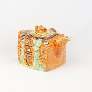 Può includere: Una teiera in ceramica marrone e verde a forma di casa con tetto di paglia e finestre. La teiera ha un beccuccio e un manico.