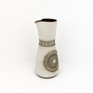 Può includere: Brocca in ceramica bianca con interno e base marrone scuro. La brocca presenta una fascia decorativa attorno al collo e un disegno circolare sul corpo. Il disegno è in un colore marrone scuro a contrasto.