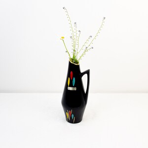 May include: A black ceramic vase with a geometric design in yellow, red, blue, and green. The vase has a narrow neck and a wide base. It is decorated with a silver label. The vase is filled with a bouquet of small white flowers and green stems.