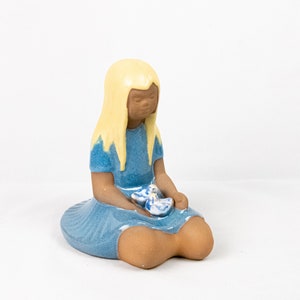 May include: A ceramic figurine of a seated girl with blonde hair and a blue dress. The girl is holding a small, blue and white object. The figurine has a light brown skin tone and is set against a white background.